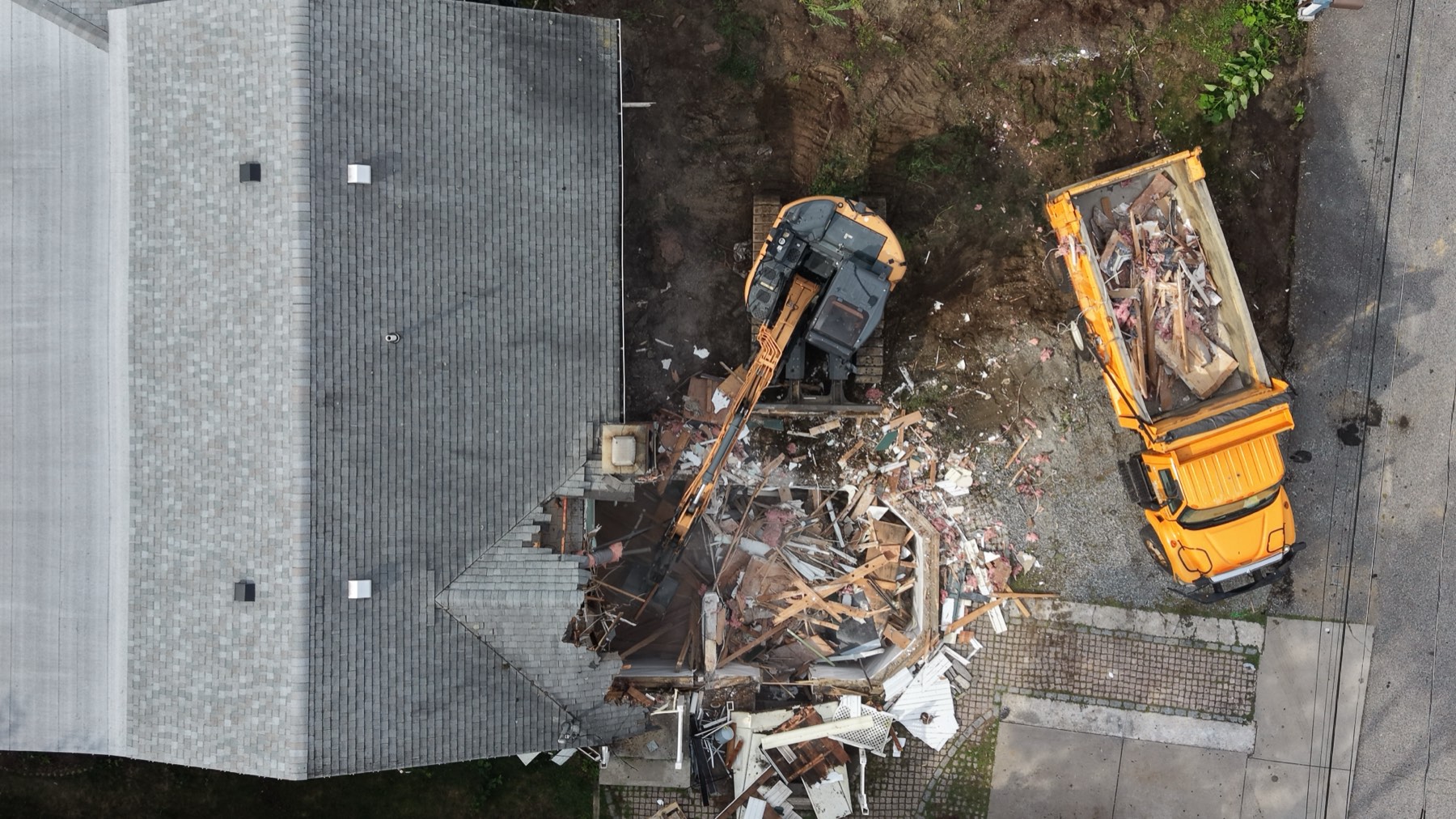 Overhead view of a roof tear-off in progress with an excavator and loaded dump truck