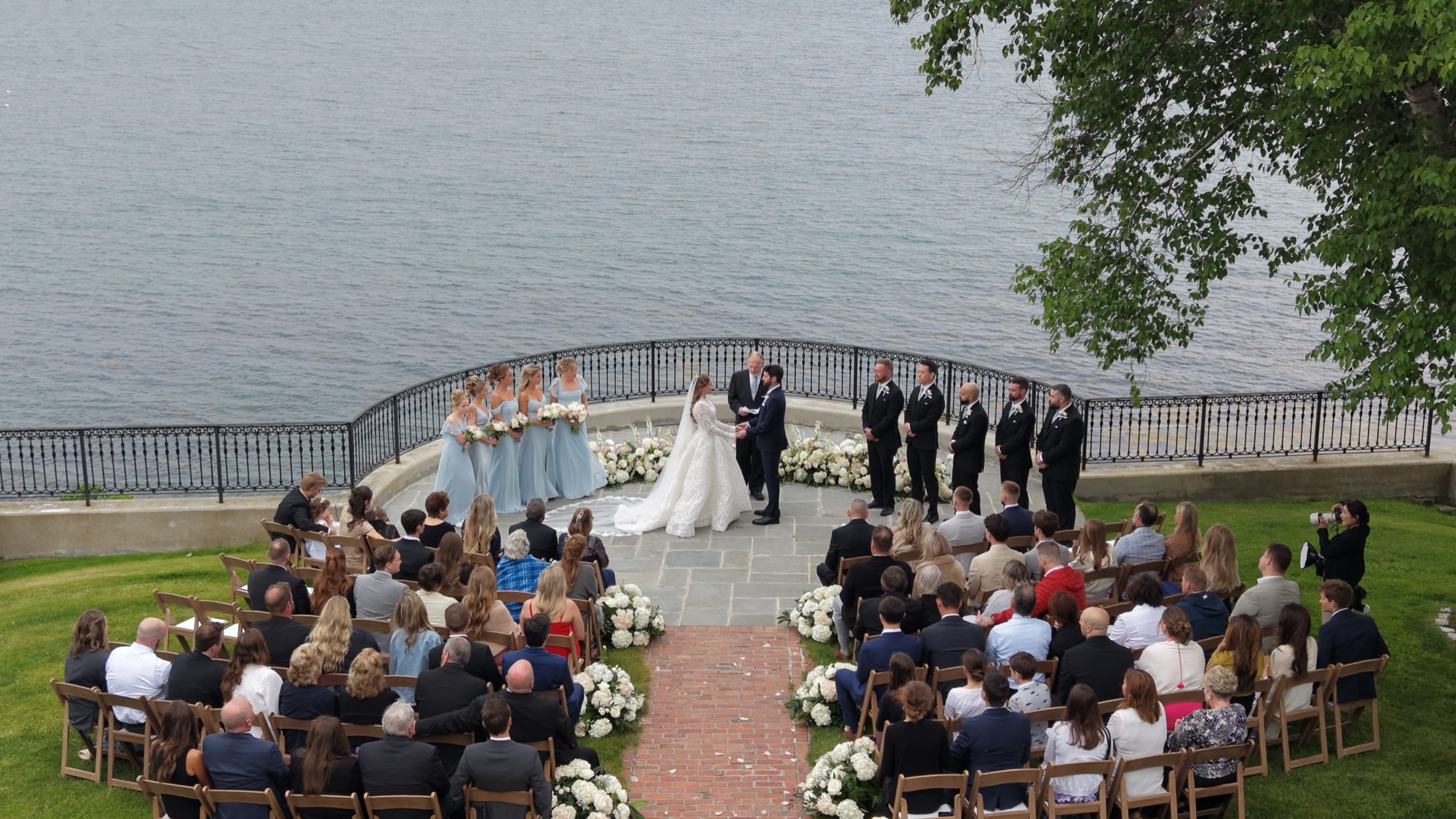 Aerial view of a waterfront wedding ceremony, guests seated along the aisle with the lake beyond